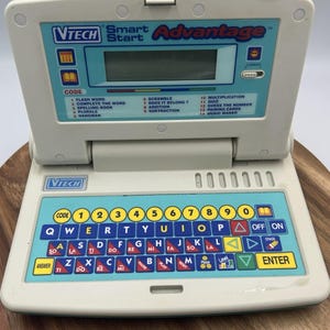 Può includere: Un giocattolo educativo elettronico VTech Smart Start Advantage con custodia in plastica grigio chiaro e pannello di controllo blu. Il dispositivo è dotato di uno schermo LCD monocromatico, una tastiera QWERTY e 14 giochi educativi stampati.