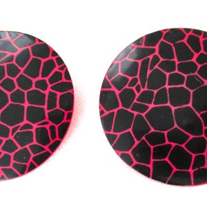 May include: Pair of round earrings with a black background and a red, irregular pattern. The earrings have a glossy finish and a simple, modern design. The earrings are a bold accessory.