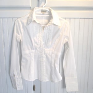 May include: A white button-down shirt with long sleeves, a collared neckline, and a fitted waist. The shirt is hanging on a white hanger against a white cabinet. The shirt has a pleated design on the front.