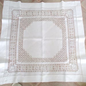 May include: A white square tablecloth with a detailed, openwork border. The border features a repeating pattern of floral and geometric shapes in a light brown color. The tablecloth is folded and laid flat, showcasing the intricate design.