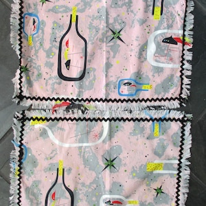 Puede incluir: Dos manteles individuales rosas con un diseño retro con contornos de botellas negras, explosiones de estrellas y formas abstractas en amarillo, rojo y azul. Los manteles individuales tienen un fondo moteado gris y blanco y están ribeteados con costuras en zigzag negras y flecos blancos.