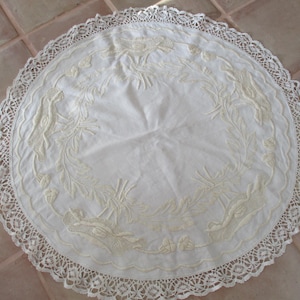 May include: A round, off-white tablecloth with intricate floral embroidery and a delicate lace edge. The embroidered design features leaves and birds. The tablecloth is made of a soft, light fabric.