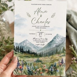 May include: A rectangular wedding invitation card featuring a watercolor illustration of a mountain landscape with a wildflower meadow in the foreground. The text reads: Together with their families, Alina & Charles, invite you to celebrate their wedding, Saturday September 27 2025 at 4:00 PM, Whispering Pines Lodge, 123 Mountain View Road, Banff, Alberta, reception to follow.
