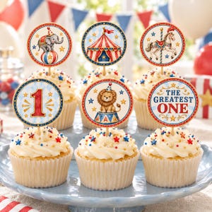 May include: Six cupcakes topped with circular circus-themed paper toppers featuring an elephant on a ball, a red and white striped tent, a carousel horse, the number 1, a lion on a pedestal, and the text THE GREATEST ONE with red and blue accents.