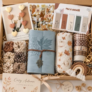 May include: A gift box containing a blue journal with a leaf print, a pencil case with autumn motifs, six patterned washi tape rolls, six fabric hair scrunchies, a set of magnetic bookmarks, heart-shaped pins, and a small macrame rainbow keychain.