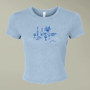Puede incluir: Una camiseta de manga corta azul claro con un gráfico de líneas azul oscuro que muestra una mesa con una vela, una botella, flores, copas, pan y un plato pequeño. El texto "girl dinner" aparece impreso en letra cursiva debajo.