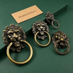 May include: Antique brass lion head door knockers with ring handles. The lion heads are detailed with manes and facial features. A small rectangular card with the text "MARIEKENLEY" is in the background. The knockers are on a dark green surface.
