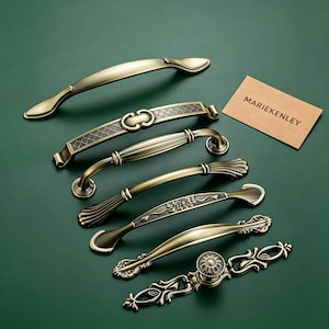 May include: A collection of six antique brass-toned metal cabinet pulls and handles with various decorative shapes, including smooth, ribbed, and floral-etched designs, accompanied by a small rectangular card printed with the text MARIEKENLEY.