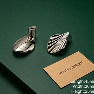 May include: Two silver-toned metal cabinet knobs with a shell-inspired design. One knob shows the mounting post, while the other displays the ridged surface. A tan card with the text MARIEKENLEY is present, noting dimensions of 45mm by 30mm by 25mm.