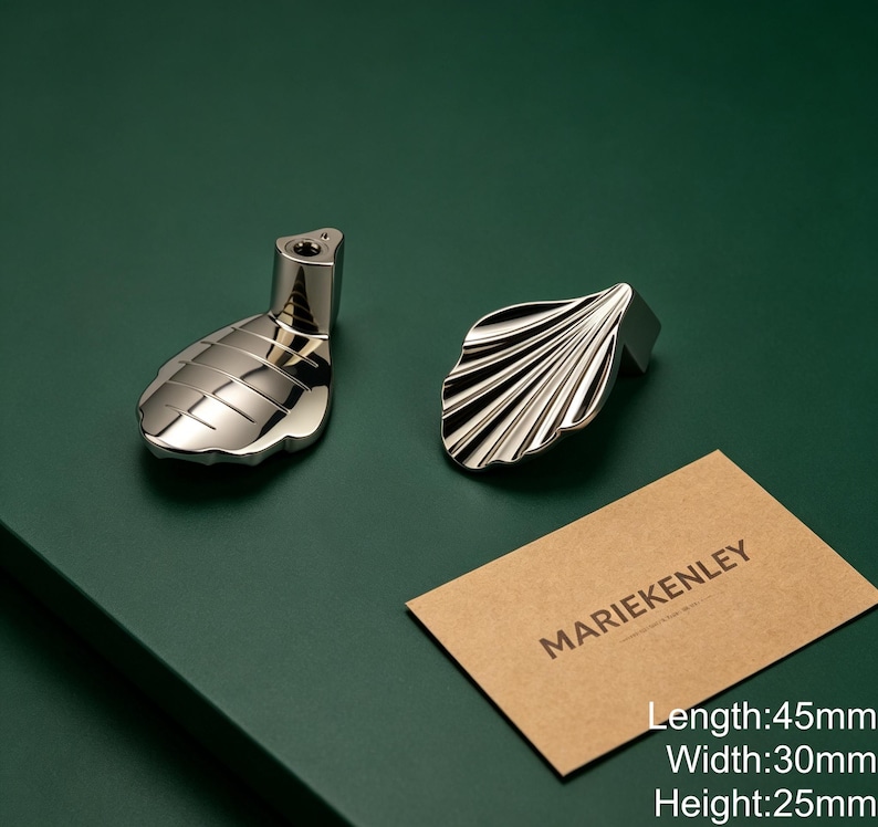 May include: Two silver-toned metal cabinet knobs with a shell-inspired design. One knob shows the mounting post, while the other displays the ridged surface. A tan card with the text MARIEKENLEY is present, noting dimensions of 45mm by 30mm by 25mm.