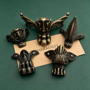 Può includere: Cinque piedini per mobili in metallo color ottone antico con design ornati a forma di artiglio e fori di montaggio. I pezzi sono disposti su un cartoncino color cuoio con il testo MARIEKENLEY. Ogni piedino presenta un rilievo dettagliato.