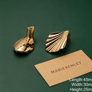 May include: Two gold-colored metal cabinet knobs shaped like shells with a polished finish. One knob shows the mounting post, while the other displays the ridged shell design. A tan card with the text MARIEKENLEY is present, noting 45mm length, 30mm width, and 25mm height.