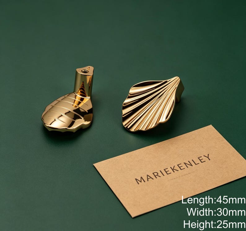 May include: Two gold-colored metal cabinet knobs shaped like shells with a polished finish. One knob shows the mounting post, while the other displays the ridged shell design. A tan card with the text MARIEKENLEY is present, noting 45mm length, 30mm width, and 25mm height.