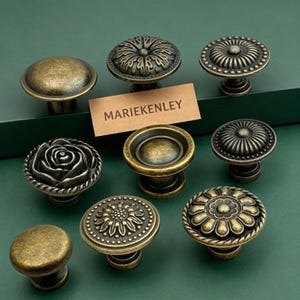 Puede incluir: Una colección de ocho pomos redondos de metal para armarios con acabado en latón antiguo. Los pomos presentan varios diseños en relieve, incluyendo motivos florales, formas de rosa y superficies lisas. Una etiqueta beige muestra el texto MARIEKENLEY.