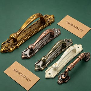 May include: Five metal door handles with ornate, decorative backplates in various finishes including antique gold, copper, bronze, cream, and a two-tone copper and bronze. Each handle features intricate embossed detailing and two mounting holes.