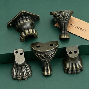 May include: Five antique brass-toned metal furniture feet with varied decorative designs, including claw-like shapes and ornate patterns with screw holes for mounting. A small tan rectangular card with the text MARIEKENLEY is included.