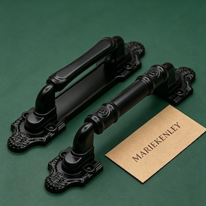 Può includere: Due maniglie per porta nero opaco con piastre di montaggio ornate e dettagliate caratterizzate da intricati motivi a volute. Una maniglia ha un'impugnatura liscia, mentre l'altra presenta anelli decorativi. Una carta beige con il testo MARIEKENLEY.