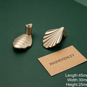 May include: Two gold-toned metal cabinet knobs with a shell-like design. One knob shows a smooth, ridged surface, while the other displays a fan-like texture. A tan card with the text MARIEKENLEY is present, noting dimensions of 45mm by 30mm by 25mm.