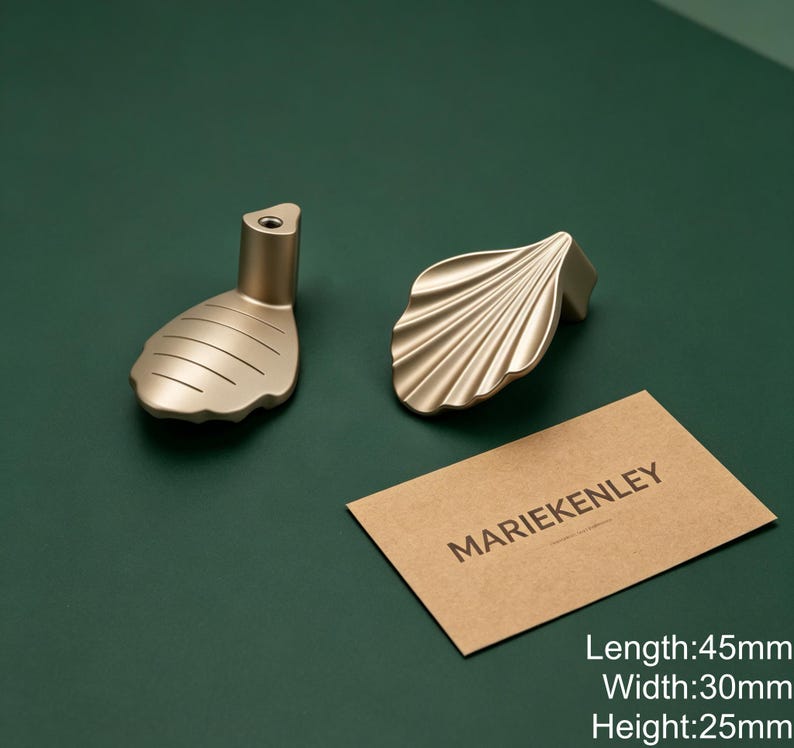 May include: Two gold-toned metal cabinet knobs with a shell-like design. One knob shows a smooth, ridged surface, while the other displays a fan-like texture. A tan card with the text MARIEKENLEY is present, noting dimensions of 45mm by 30mm by 25mm.