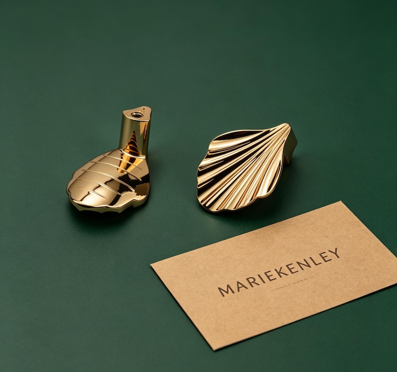 May include: Two gold-colored metal cabinet knobs with a polished finish. One knob features a smooth, rounded shape with horizontal lines, while the other has a fan-like, scalloped design. A rectangular tan card displays the text MARIEKENLEY.