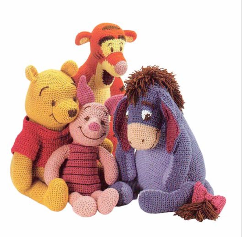 May include: A group of four crocheted plush toys featuring characters from Winnie the Pooh. The toys include Winnie the Pooh in yellow, Piglet in pink, Tigger in orange, and Eeyore in blue. Each toy has distinct features.