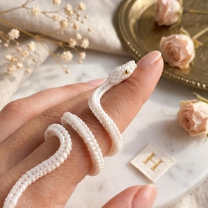 May include: A white beaded snake ring crafted from small, uniform seed beads. The snake features a single gold-toned bead for an eye and is coiled in a spiral shape, designed to wrap around a finger with a flexible, textured beaded body.