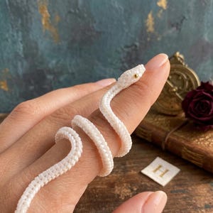 May include: A white beaded snake ring crafted from small, uniform beads. The snake features a single gold-colored bead for an eye and is coiled in a spiral shape, designed to wrap around a finger with a textured, woven appearance.