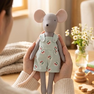 May include: A gray mouse doll with large pink ears and a long tail. The doll wears a light green romper with a cherry pattern. The doll is being held in someone's hands. The background includes a vase of flowers and wooden toys.