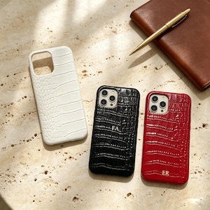 May include: Three smartphone cases with a crocodile-embossed texture in white, black, and red. Each case features a square camera cutout and gold-colored monogrammed initials. The white case has initials, the black case has FA, and the red case has ER.