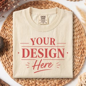 Könnte beinhalten: Ein cremefarbenes Kurzarm-T-Shirt mit Rundhalsausschnitt. Auf der Vorderseite befindet sich ein roter Aufdruck mit dem Text YOUR DESIGN Here, der durch horizontale Linien und kleine dekorative Striche ergänzt wird. Ein Comfort Colors Etikett ist sichtbar.