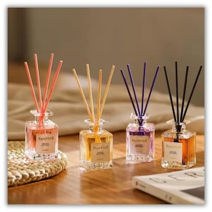 May include: Four square glass reed diffuser bottles with FADOYVUE labels, each containing coloured liquid and matching reed sticks in coral, yellow, purple, and black, arranged in a row on a wooden surface with a white book nearby.