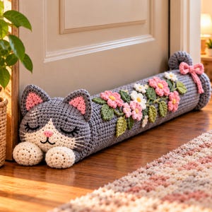 May include: A gray and white crocheted cat draft stopper featuring a sleeping face with pink inner ears and nose. The long cylindrical body is decorated with crocheted pink and white flowers and green leaves, finished with a pink bow at the end.