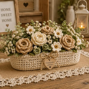 Könnte beinhalten: Ein ovaler Häkelkorb aus cremefarbenem Garn, gefüllt mit gehäkelten Blumen in Beige-, Creme- und Grüntönen. Ein kleines hölzernes Herzschild, das mit einer Schnur befestigt ist, trägt den Text "handmade with love" und ein kleines Herzsymbol.