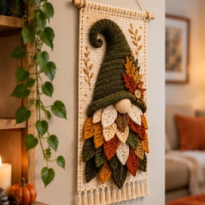 Op de afbeelding: Een rechthoekige cr&egrave;mekleurige gehaakte wandhanger met een kabouter met een hoge olijfgroene muts en een baard gemaakt van gelaagde, veelkleurige gehaakte blaadjes in tinten oranje, geel, bruin en groen, afgewerkt met een houten knoop.