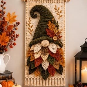 May include: A crochet wall hanging featuring a gnome with a tall olive green hat, a round beige nose, and a beard made of layered fabric leaves in shades of cream, brown, orange, and green. The gnome is attached to a cream-colored rectangular base.