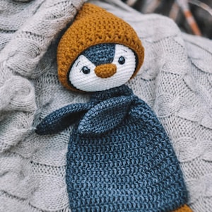 Könnte beinhalten: Eine gehäkelte Schnuffeltuch-Puppe in Form eines Pinguins mit weißem Gesicht, schwarzen Augenpartien und einem braunen Schnabel. Der Pinguin trägt eine braune Mütze und hat einen dunkelblauen Körper mit passenden braunen Füßen aus Garn.