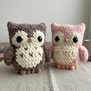 May include: Two handmade crochet owl figurines. One owl is brown and cream, the other is pink and cream. Both have large black eyes and small beaks. The owls are sitting on a light-colored surface.