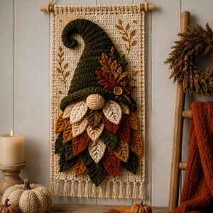 Op de afbeelding: Een wandhanger met een gehaakte kabouter met een donkergroene muts, een ronde neus en een baard van gelaagde, veelkleurige gehaakte bladeren in cr&egrave;me, oranje, bruin en groen, opgehangen aan een houten stok met een touwtje.