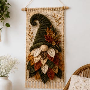 Op de afbeelding: Een wandhanger met een gehaakte kabouter met een hoge, donkergroene puntmuts en een baard van gelaagde, veelkleurige stoffen bladeren in tinten cr&egrave;me, goud, oranje en groen. De kabouter is bevestigd op een rechthoekig, cr&egrave;mekleurig macram&eacute; paneel.
