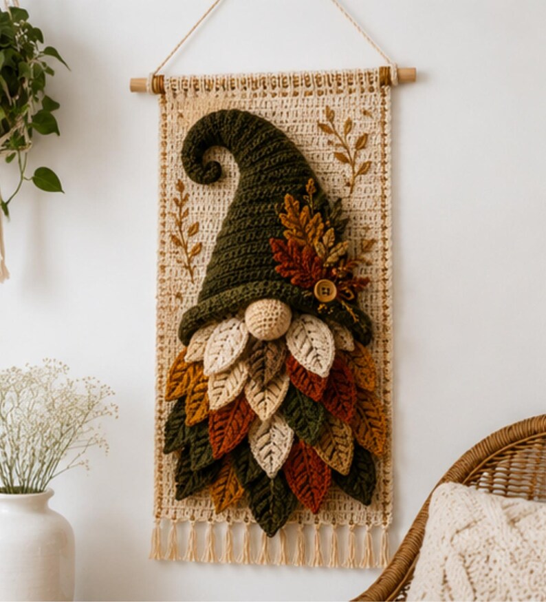 Op de afbeelding: Een wandhanger met een gehaakte kabouter met een hoge, donkergroene puntmuts en een baard van gelaagde, veelkleurige stoffen bladeren in tinten cr&egrave;me, goud, oranje en groen. De kabouter is bevestigd op een rechthoekig, cr&egrave;mekleurig macram&eacute; paneel.