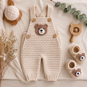 May include: A beige crochet overall featuring a central bear face appliqué on the chest, two shoulder straps, and four wooden buttons. The set includes a pair of matching beige crochet booties, each adorned with a small bear face appliqué on the front.