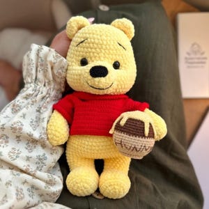 Könnte beinhalten: Ein gelbes, gehäkeltes Bären-Stofftier in einem roten T-Shirt. Der Bär hat schwarze Augen und eine schwarze Nase und hält einen kleinen braunen Honigtopf mit dem Wort HUNNY in dunklem Garn, verziert mit hellgelben Honigtropfen.