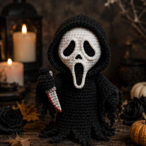May include: A crocheted figure wearing a black hooded robe and a white mask with large black eye and mouth openings. The figure holds a small gray knife with a red-tipped blade in its right hand. The robe features a frayed, uneven hemline.
