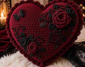 Puede incluir: Un cojín de ganchillo en forma de corazón de color burdeos intenso con borde festoneado, aplicaciones florales negras y un detalle de mariposa negra. La superficie está decorada con pequeñas cuentas negras y un dije de corazón negro.