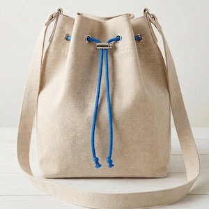 May include: A beige canvas bucket bag featuring a drawstring closure with a bright blue rope cord. The bag includes a long shoulder strap and silver-toned metal grommets through which the blue cord is threaded to secure the top opening.