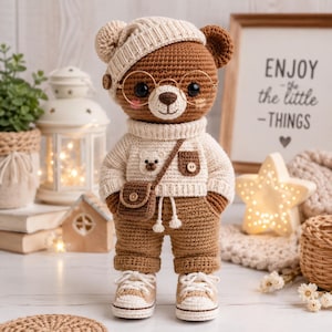 Könnte beinhalten: Ein gehäkelter brauner Teddybär mit einer cremefarbenen Mütze, einem passenden Rollkragenpullover mit kleinem Bärenaufnäher und Tasche, brauner Hose und cremefarbenen Turnschuhen. Der Bär trägt eine runde goldene Brille und eine kleine braune Umhängetasche.