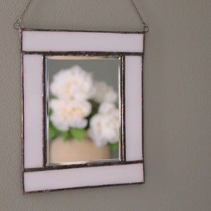 May include: A white framed mirror with a white border and a silver interior. The mirror reflects a bouquet of white flowers in a vase.