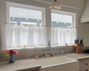 May include: White linen kitchen curtains hanging on brass rods. The curtains are draped over two windows, with a view of a light blue building and a white roof. A white farmhouse sink and a countertop with a vase of flowers are in the foreground.