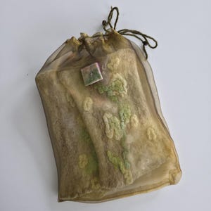 May include: A rectangular, semi-sheer, bronze-colored organza drawstring pouch containing a textured, felted wool item. The felted piece is beige with light green floral-like patterns and features a small, square, multi-colored decorative tag.