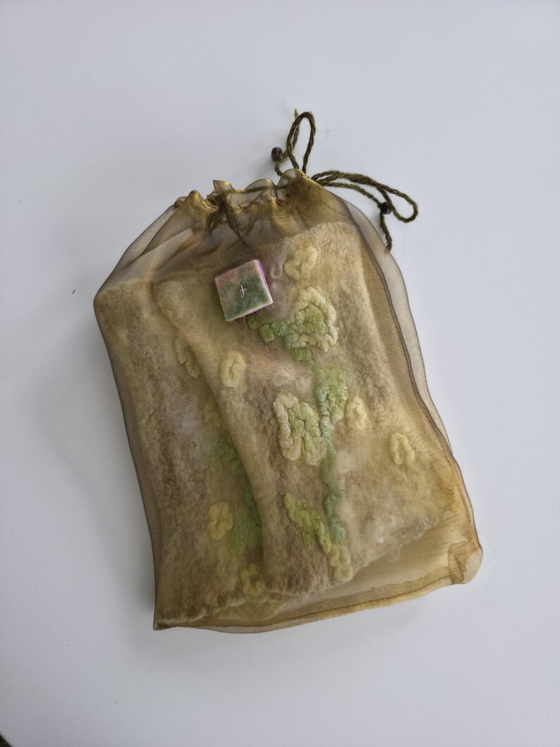 May include: A rectangular, semi-sheer, bronze-colored organza drawstring pouch containing a textured, felted wool item. The felted piece is beige with light green floral-like patterns and features a small, square, multi-colored decorative tag.
